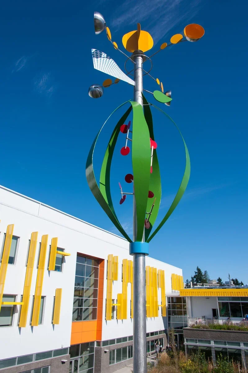 Photosynthesis Whirligig with green leaf forms, red discs and yellow wind-driven elements against a blue sky.